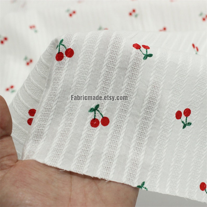 May include: White cotton fabric with red cherries and thin white stripes. The fabric has a textured surface. "Fabricmade.etsy.com" is printed on the fabric.