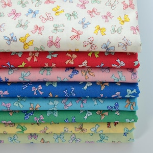 Könnte beinhalten: Ein Stapel bunter Stoffballen mit einem sich wiederholenden Schleifenmuster. Die Stoffe sind in verschiedenen Farben gehalten, darunter Rot, Rosa, Blau und Grün, mit Schleifen in passenden Farben. Der oberste Stoff ist cremefarben mit mehrfarbigen Schleifen.