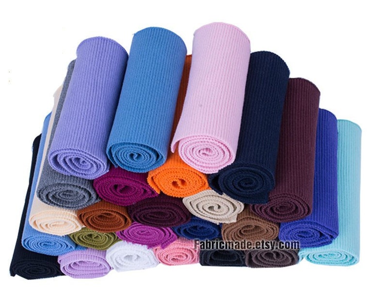 Op de afbeelding: Een verzameling van 20 rollen geribbelde breistof in verschillende kleuren, waaronder blauw, roze, oranje, bruin en grijs. De stof is gevouwen en gestapeld in een stapel. Fabricmade.etsy.com is afgedrukt op de onderkant van de stapel.