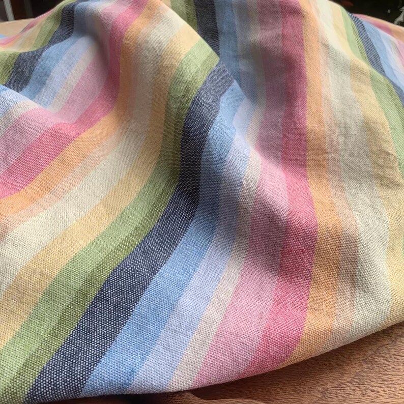 May include: A piece of fabric featuring a vertical striped pattern in various pastel and muted colors including pink, light blue, yellow, green, and dark navy blue. The textile has a visible woven texture and is draped to show folds and depth.