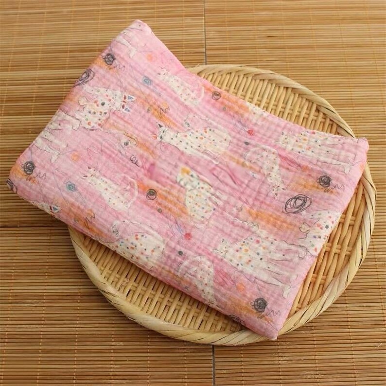 May include: A folded pink muslin blanket featuring a repeating pattern of white cats with colorful polka dots and small dark circular scribbles. The fabric has a visible crinkled texture and is layered on a round woven bamboo tray.