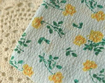 May include: A white floral patterned fabric featuring yellow roses and green leaves. The fabric has a textured surface and is partially covering a cream-colored, open-weave doily. The design is reminiscent of vintage textiles.