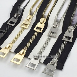 Può includere: Quattro tiretti per cerniera in diverse finiture: nero, oro, argento e nichel. Le cerniere sono disposte su un tessuto a strisce bianche e nere.