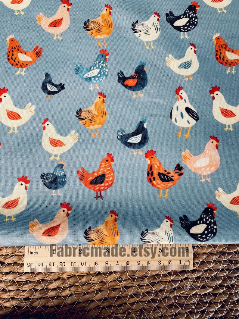 May include: A light blue fabric featuring a repeating pattern of chickens in various colors including white, orange, black, and blue. A wooden ruler is positioned at the bottom, showing measurements in inches and centimeters for scale.