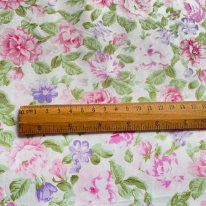 May include: White fabric with a pink and purple floral print. A wooden ruler with centimetre markings is placed on top of the fabric.