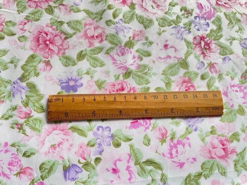 May include: White fabric with a pink and purple floral print. A wooden ruler with centimetre markings is placed on top of the fabric.