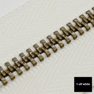 May include: Close-up of a zipper with antique gold metal teeth on a white fabric background. The text "1 off white" is visible in the lower right corner of the image.
