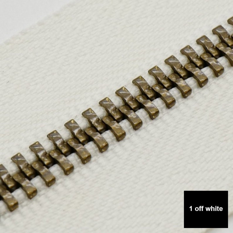 May include: Close-up of a zipper with antique gold metal teeth on a white fabric background. The text "1 off white" is visible in the lower right corner of the image.
