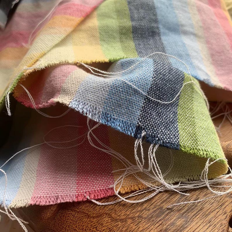 May include: A piece of woven fabric featuring a vertical striped pattern in pastel shades of pink, yellow, light blue, and green. The textile has a visible textured weave and frayed edges with loose white threads extending from the cut ends.