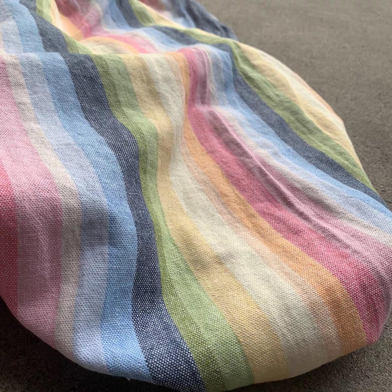 May include: A textile fabric featuring a repeating vertical striped pattern in a palette of soft pastel colors including pink, light blue, yellow, green, and cream. The material has a visible woven texture with a consistent weave throughout the piece.
