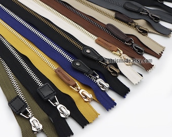 May include: Assortment of zipper tapes in various colors, including black, brown, beige, yellow, and blue. The zippers have metal teeth and are arranged in a fan shape. The zipper pulls are made of metal and leather.
