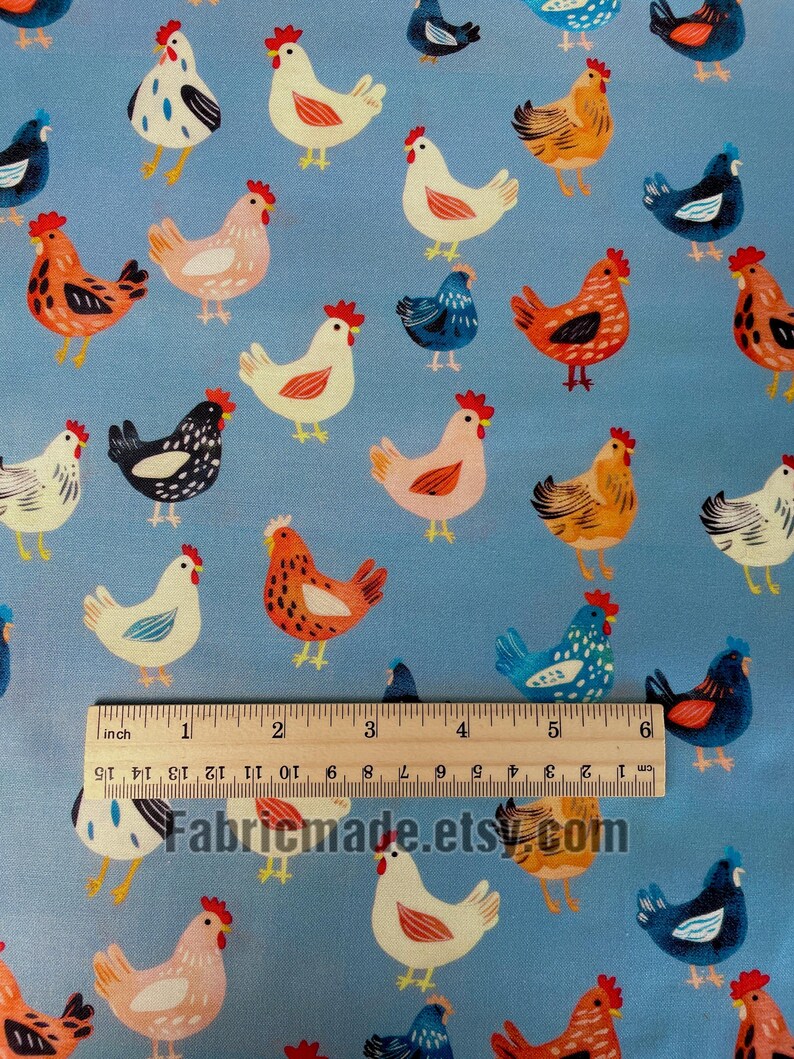 May include: A light blue fabric featuring a repeating pattern of chickens in various colors including white, orange, and dark blue. A wooden ruler is placed across the fabric, showing a measurement scale of 6 inches and 15 centimeters.