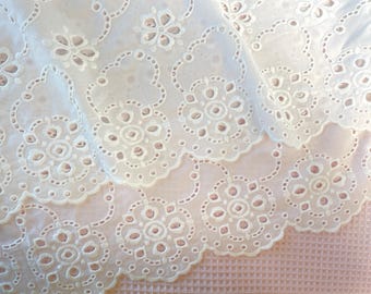 May include: Close-up of white eyelet fabric with a floral and scalloped edge design. The fabric is layered, revealing a glimpse of a pink textured surface underneath. This delicate fabric is suitable for sewing and crafting projects.