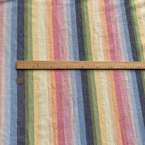 May include: A woven fabric featuring vertical stripes in shades of blue, green, yellow, orange, and pink. A wooden ruler is placed horizontally across the fabric, measuring approximately 16.5 inches in length, alongside a small circular metal coin.