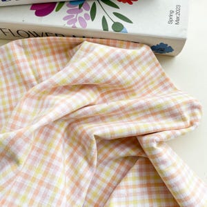 May include: Close-up of a fabric with a gingham pattern in shades of peach, yellow, and white. The fabric is draped, showing the texture and weave of the material. The fabric is likely cotton or linen.