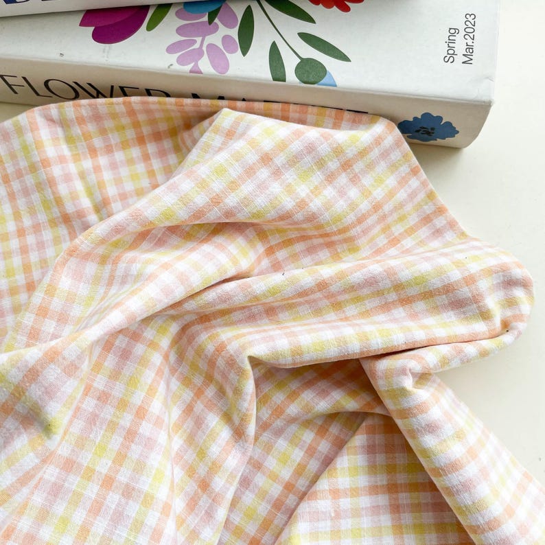 May include: Close-up of a fabric with a gingham pattern in shades of peach, yellow, and white. The fabric is draped, showing the texture and weave of the material. The fabric is likely cotton or linen.