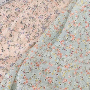 Puede incluir: Dos piezas de tela con estampado floral. La pieza superior es una tela rosa claro con un pequeño estampado floral y la pieza inferior es una tela azul claro con un estampado floral más grande. Ambas telas tienen un delicado patrón de ojales.