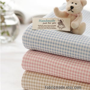 Peut inclure: Pile de trois échantillons de tissu vichy en bleu, rose et beige. Le tissu a un petit motif à carreaux. Un petit ours en peluche est assis sur le dessus du tissu.