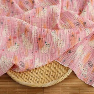 May include: A soft, crinkled pink fabric featuring a repeating pattern of white cats with orange and black spots, interspersed with small orange and black circular scribbles, draped over a round, light-colored woven bamboo tray.