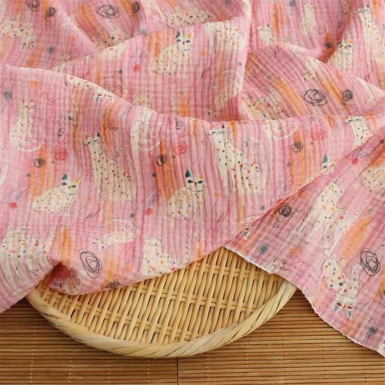 May include: A soft, crinkled pink fabric featuring a repeating pattern of white cats with orange and black spots, interspersed with small orange and black circular scribbles, draped over a round, light-colored woven bamboo tray.
