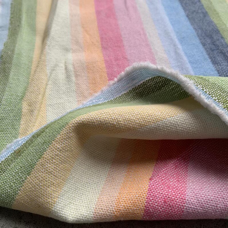 May include: A woven fabric featuring vertical stripes in a pastel color palette of light green, yellow, peach, pink, light blue, and grey. The textile has a visible textured weave and a frayed edge where the material is folded over itself.