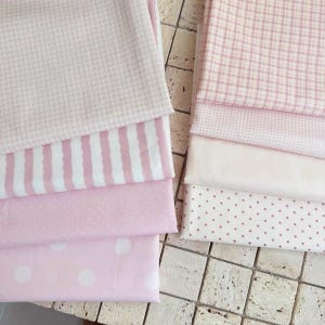 May include: A collection of folded fabric pieces in various shades of pink and white. The patterns include a fine grid, vertical stripes, small polka dots, and large white polka dots on a light pink background, all stacked neatly in two columns.