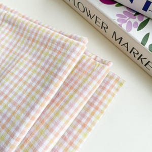 May include: A stack of four pastel-colored gingham fabric napkins. The napkins are in shades of pink, yellow, and orange, with a white background. The text "FLOWER MARKET" is partially visible in the background.
