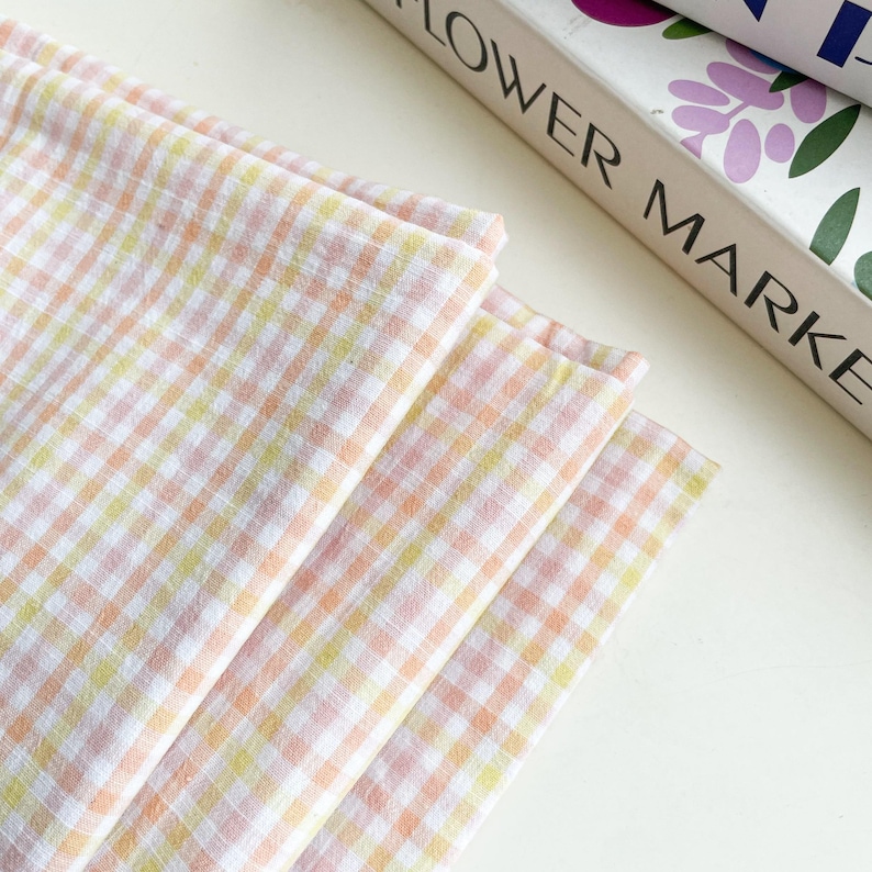 May include: A stack of four pastel-colored gingham fabric napkins. The napkins are in shades of pink, yellow, and orange, with a white background. The text "FLOWER MARKET" is partially visible in the background.
