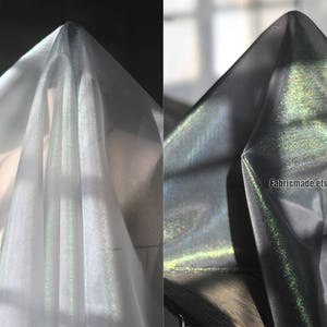May include: Two images of iridescent fabric. The fabric is draped, creating folds and shadows. The fabric appears to shift colors, with hints of green and silver. The fabric is semi-transparent, allowing light to pass through. The Etsy shop name "Fabricmade.etsy.com" is visible.