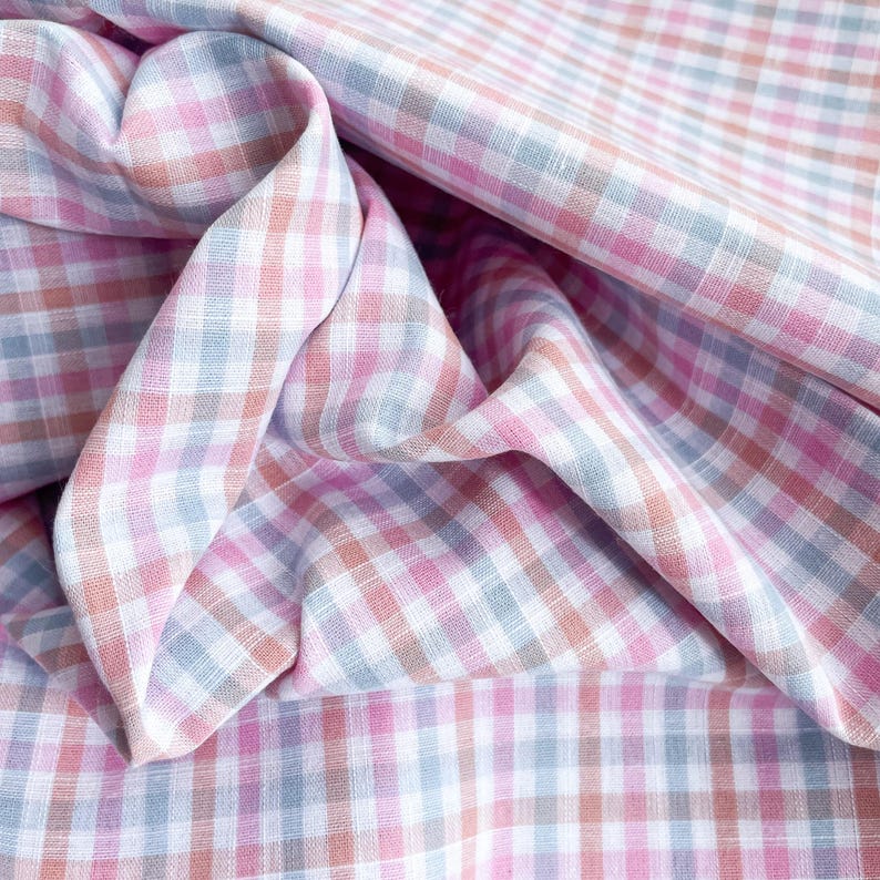 May include: Close-up of a fabric with a gingham pattern in shades of pink, white, gray, and peach. The fabric is folded and draped, showing the texture and pattern variations. The overall theme is textile design.