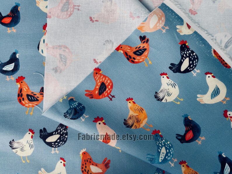 May include: A blue fabric featuring a repeating pattern of chickens in various colors including orange, black, white, and blue. The fabric is folded to reveal a lighter, off-white underside. The text Fabricmade.etsy.com is printed on the fabric.