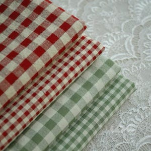 Peut inclure: Trois carrés de tissu pliés avec des motifs vichy rouges, blancs et verts. Les tissus sont empilés en diagonale, mettant en valeur les différentes combinaisons de couleurs. Le fond est un tissu en dentelle blanche.