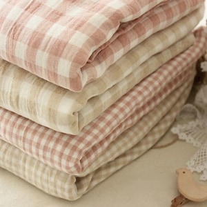Puede incluir: Una pila de piezas de tela dobladas con un patrón de cuadros vichy. Los textiles alternan entre una combinación de colores rosa empolvado suave y blanco y una combinación neutra de beige y blanco, mostrando un tejido ligero y texturizado.
