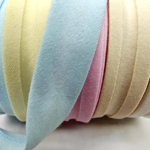 May include: Close-up of a roll of bias tape in pastel colors. The tape is folded over, revealing a light blue, yellow, pink, and beige fabric. The fabric has a subtle texture and is tightly wound.
