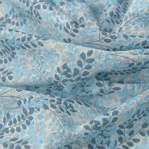 May include: Close-up of a fabric with a delicate floral pattern. The fabric features a repeating design of blue and grey leaves and small floral elements on a white background. The fabric has a soft, textured appearance.