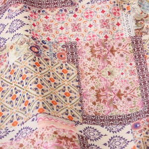 Op de afbeelding: Close-up van een stof met een patchwork-ontwerp. De stof heeft een mix van bloemen- en geometrische patronen in tinten roze, paars, oranje en crème. Het ontwerp bevat ingewikkelde randen en een verscheidenheid aan texturen.