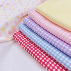 Puede incluir: Una pila de tela de cuadros de colores en tonos rojo, azul, morado, rosa y amarillo. La tela está doblada cuidadosamente, mostrando el clásico patrón de cuadros. En el fondo se ve una tela blanca con lunares pastel.