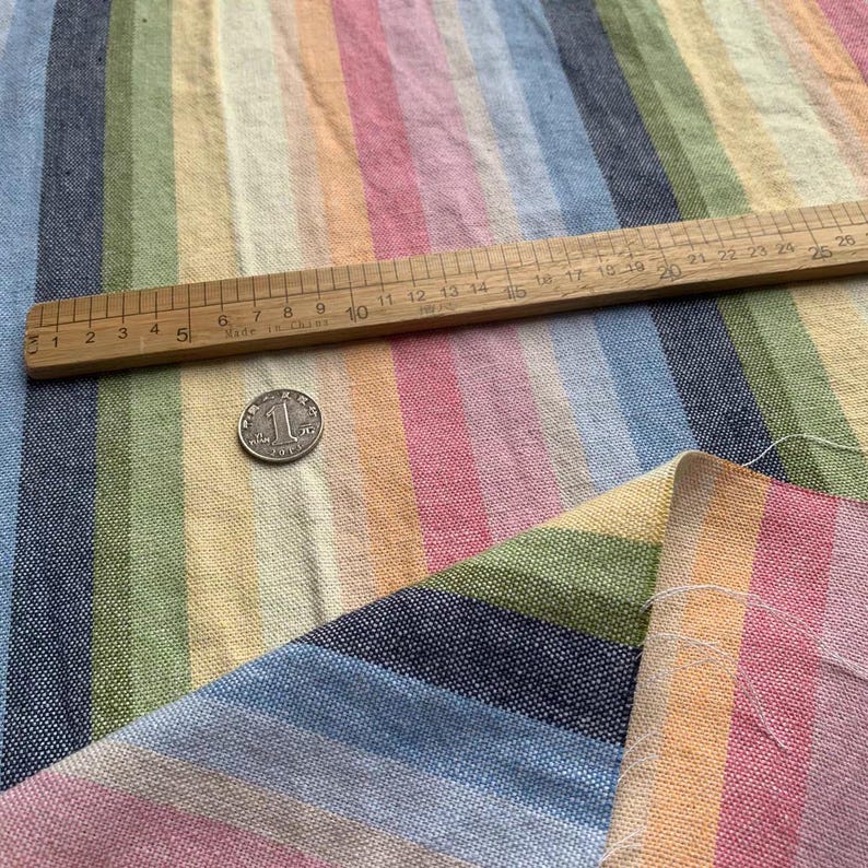 May include: A woven fabric featuring vertical stripes in shades of blue, green, yellow, and pink. A wooden ruler is placed across the fabric, showing measurements in inches, and a silver coin is positioned on the surface for scale reference.