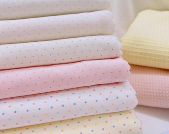 May include: A stack of pastel-colored fabric rolls, each adorned with small, colorful polka dots. The colors include pink, yellow, and blue, creating a soft and inviting aesthetic. The fabric appears to be soft and suitable for baby items.