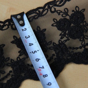 May include: Close-up of black floral lace with a blue measuring tape. The lace features a delicate, embroidered design with flowers and leaves. The tape measure shows a measurement of approximately 7.6 cm.