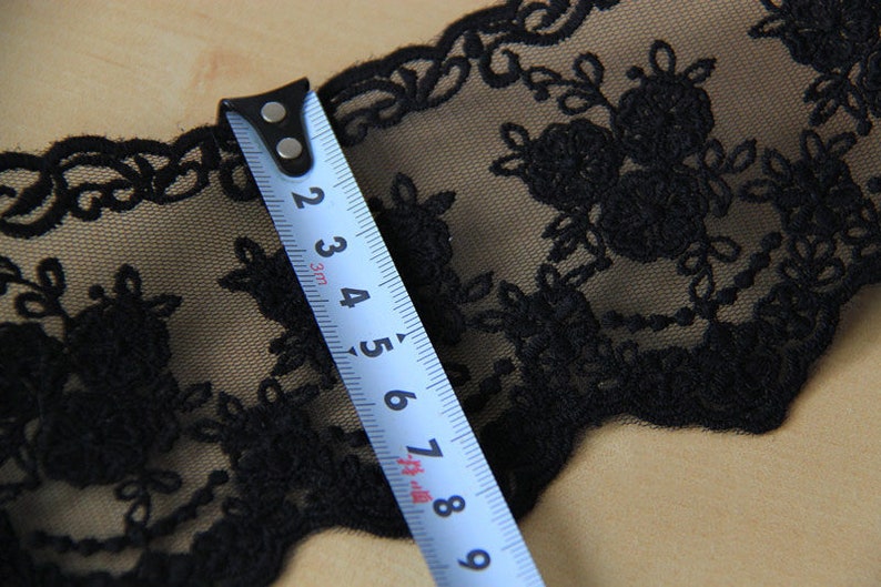 May include: Close-up of black floral lace with a blue measuring tape. The lace features a delicate, embroidered design with flowers and leaves. The tape measure shows a measurement of approximately 7.6 cm.