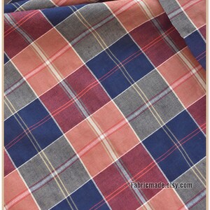 May include: Red, blue, and gray plaid fabric with a white stripe. The fabric is folded and has a slight wrinkle. The text "Fabricmade.etsy.com" is in the bottom right corner.