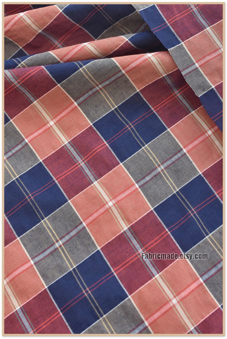 May include: Red, blue, and gray plaid fabric with a white stripe. The fabric is folded and has a slight wrinkle. The text "Fabricmade.etsy.com" is in the bottom right corner.