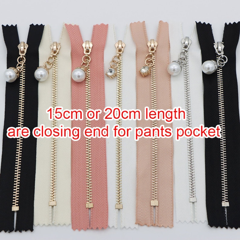 May include: Assortment of seven zipper closures with gold metal teeth and different colored tapes. The zippers have decorative pulls with a pearl and a rhinestone. The text "15cm or 20cm length are closing end for pants pocket" is printed below the zippers.