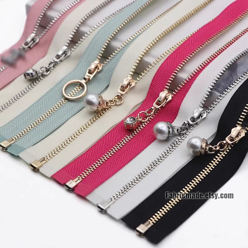 May include: Assortment of zipper tapes with different colors and finishes, including gold, silver, and black. Each zipper tape has a decorative pull with a pearl or rhinestone accent. The zipper tapes are arranged in a row, with the gold zipper tape at the bottom.