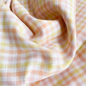 May include: Close-up of a folded fabric with a gingham pattern. The fabric features a repeating pattern of white, yellow, and peach-colored squares. The fabric is soft and appears to be lightweight, with visible texture and folds.