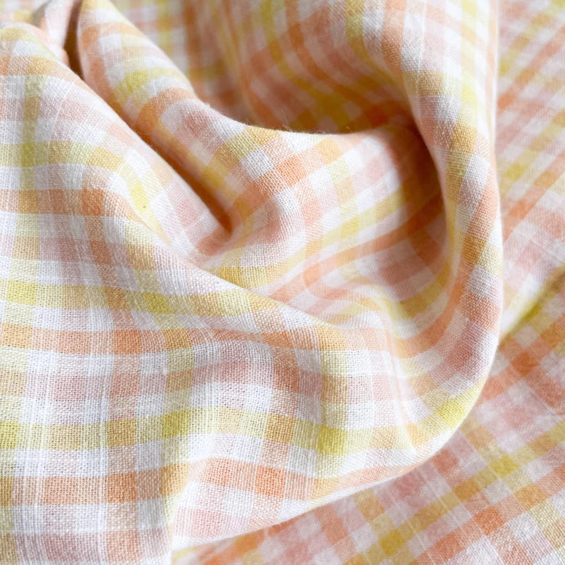 May include: Close-up of a folded fabric with a gingham pattern. The fabric features a repeating pattern of white, yellow, and peach-colored squares. The fabric is soft and appears to be lightweight, with visible texture and folds.