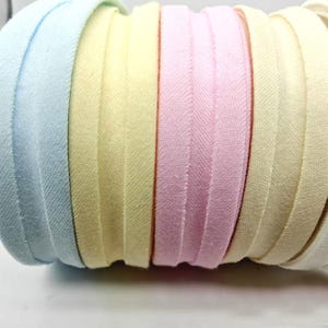May include: Close-up of a roll of bias tape in pastel colors. The tape is made of fabric and features a light blue, yellow, pink, and cream color scheme. The edges are neatly folded and stitched, creating a clean, finished look.