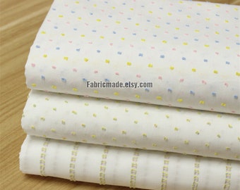May include: Three pieces of white fabric with different patterns. The top piece has a small blue, pink, and yellow dot pattern. The middle piece has a small yellow dot pattern. The bottom piece has a thin yellow stripe pattern. The fabric is folded and stacked on a wooden surface. Fabricmade.etsy.com is printed on the top piece of fabric.