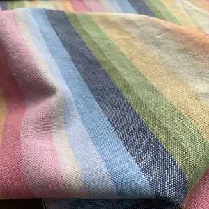 May include: A woven fabric featuring a repeating pattern of vertical stripes in various colors including pink, light blue, dark blue, sage green, and pale yellow. The textile has a visible textured weave and is folded to show depth and dimension.